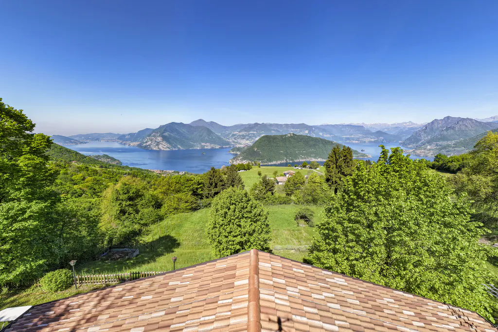 View from a terracotta roof overlooking green hills, a blue lake, and mountains under a clear blue sky.