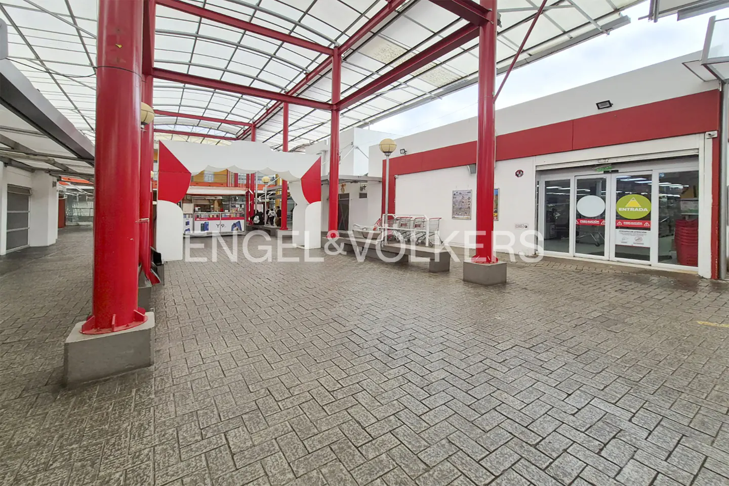 Exterior view of a shopping center with red steel beams and a brick-patterned walkway. Entrance doors are visible.