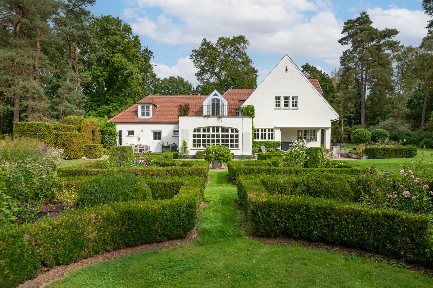 White house with red tile roof surrounded by green manicured hedges and lawn under a blue sky with white clouds.
