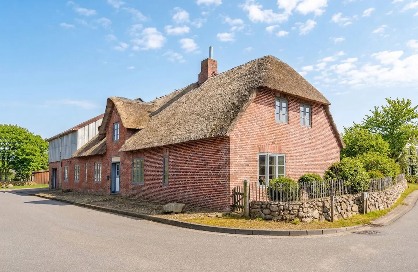 A red brick house with a thatched roof on a sunny day. The house has gray window frames and a stone wall.