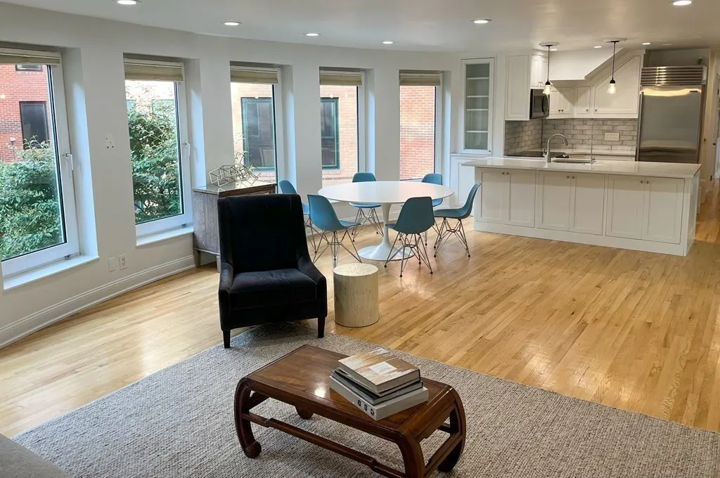 Bright, open-concept living space with hardwood floors, a white kitchen, and a dining area with blue chairs. A dark armchair sits near a coffee table.