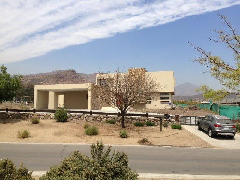 Beige modern house with stone accents, a bare tree in front, and mountains in the background under a blue sky. A gray car sits in the driveway.