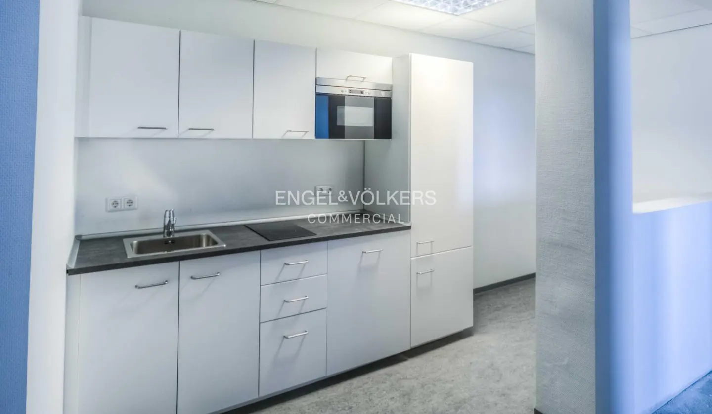 A modern, white office kitchen with stainless steel sink, black countertop, and built-in microwave. The Engel & Völkers Commercial logo is visible.