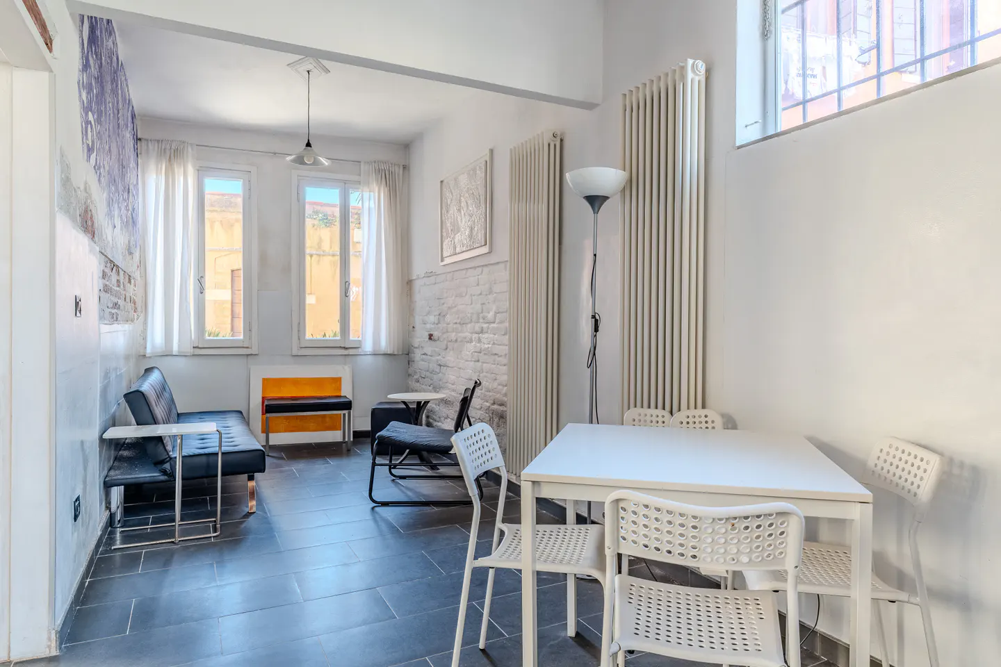 Bright, airy living space with white walls, black tile floor, and a white table with four chairs. A black sofa sits near windows with white curtains.