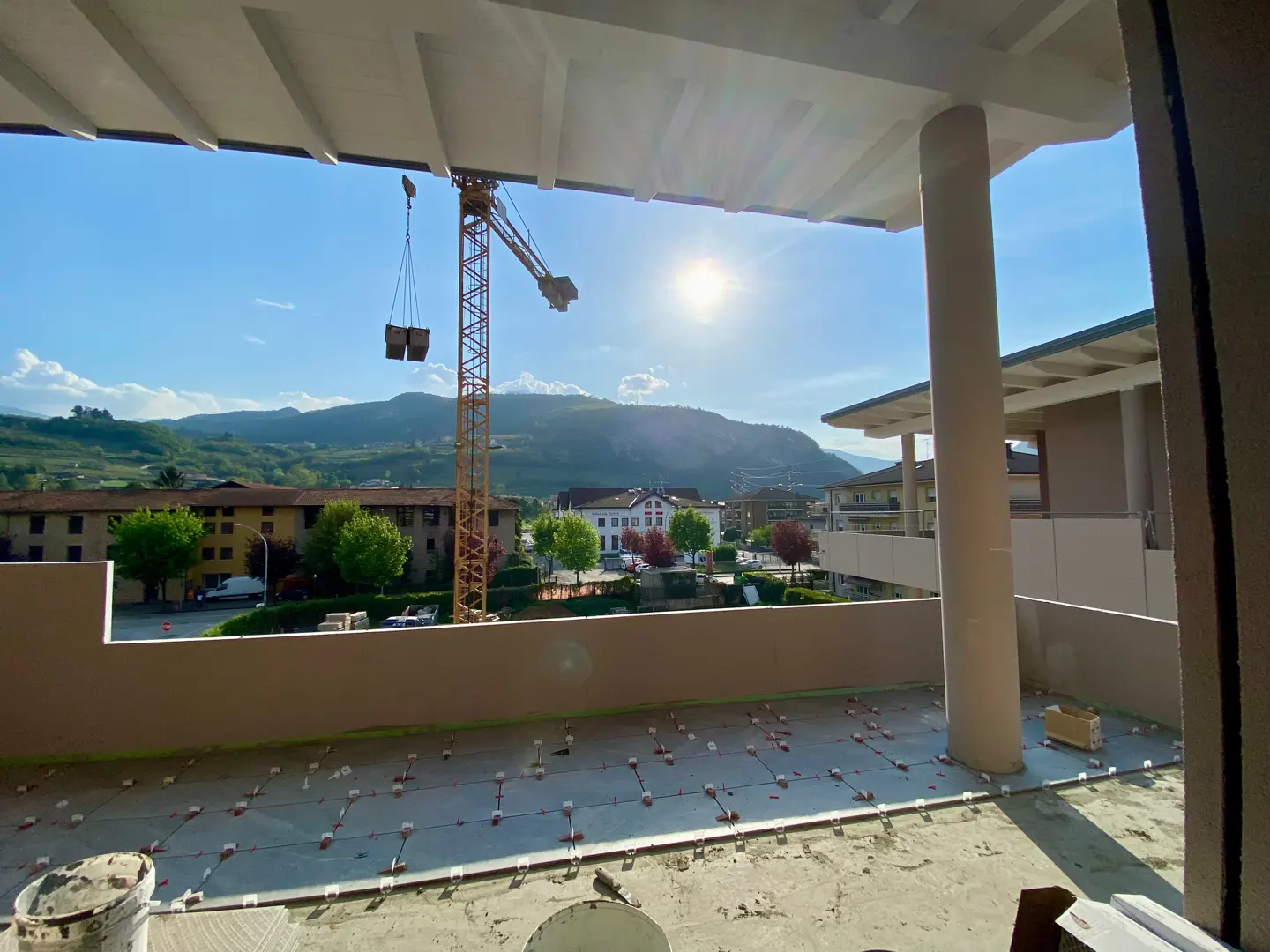 Balcony view of a construction site with a crane lifting materials, set against a backdrop of mountains and a sunny sky.