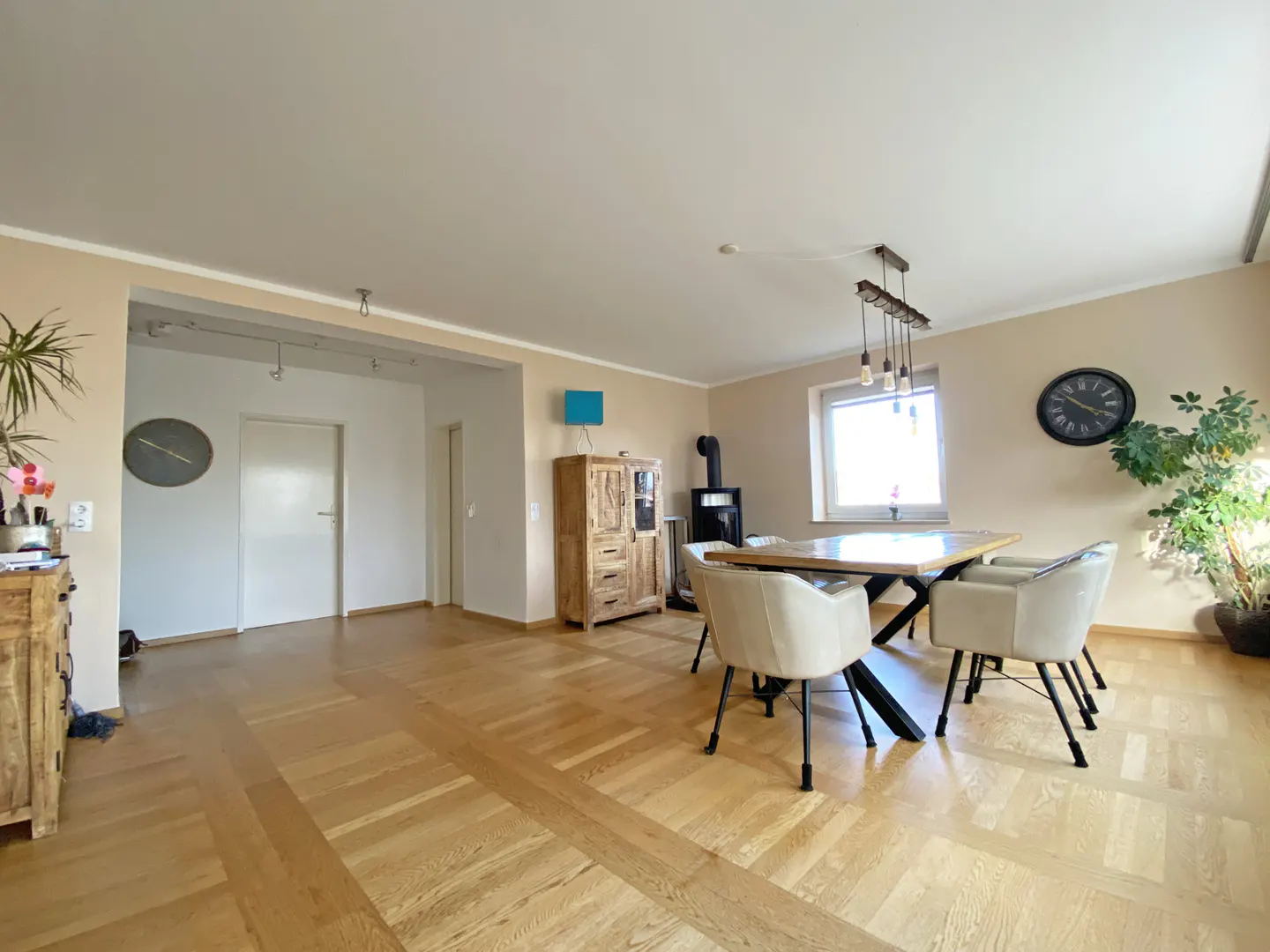 A bright, open-plan living and dining area with wood floors, a dining table with four chairs, and a wood-burning stove.