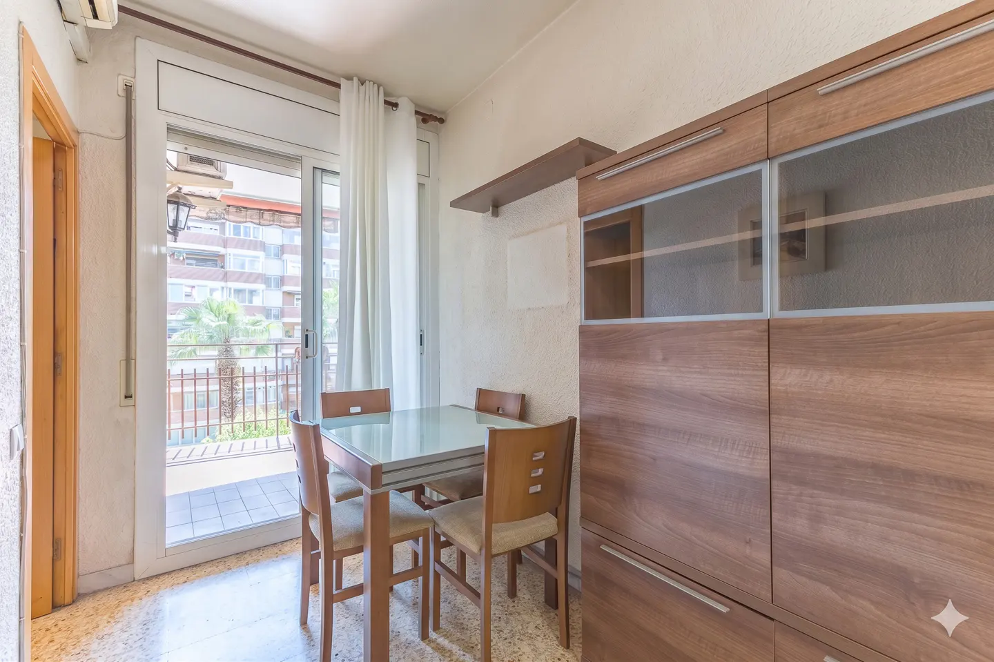 A dining area with a glass-topped table and four chairs, next to a large wooden cabinet with glass doors and a balcony view.