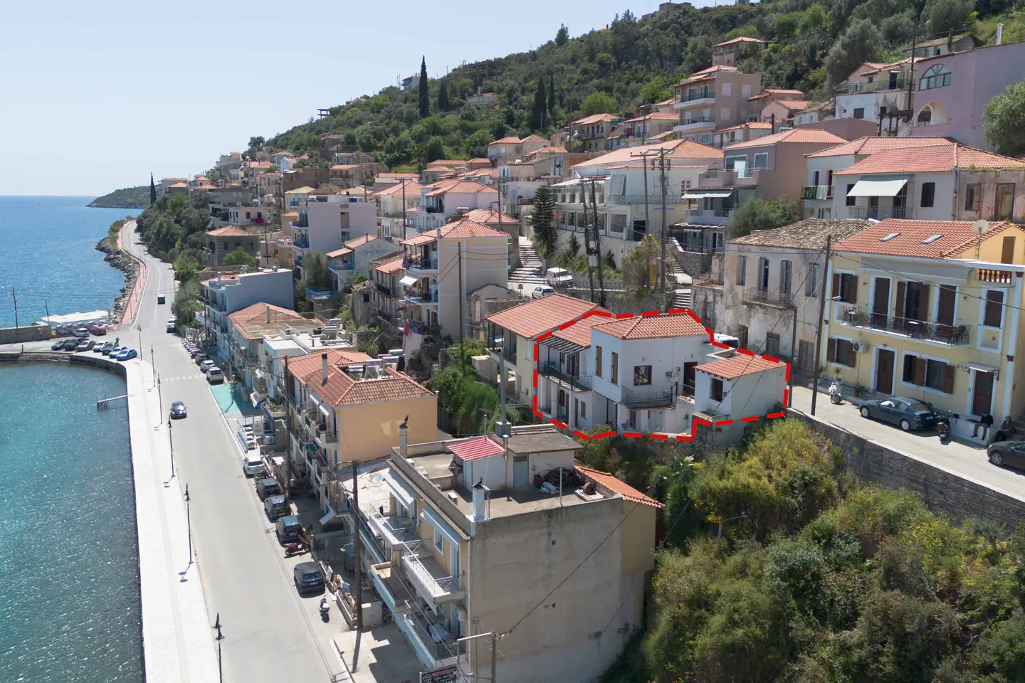 Aerial view of a white house with a red roof, outlined in red, in a coastal village with a harbor and hillside homes.