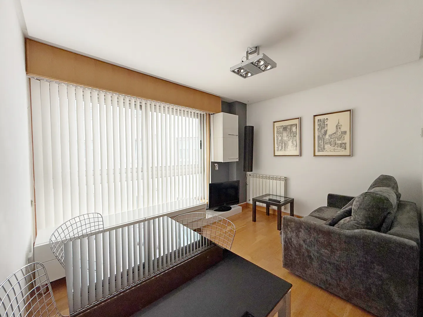 Living room with a gray sofa, black table, and white vertical blinds. Two framed pictures hang on the white wall.