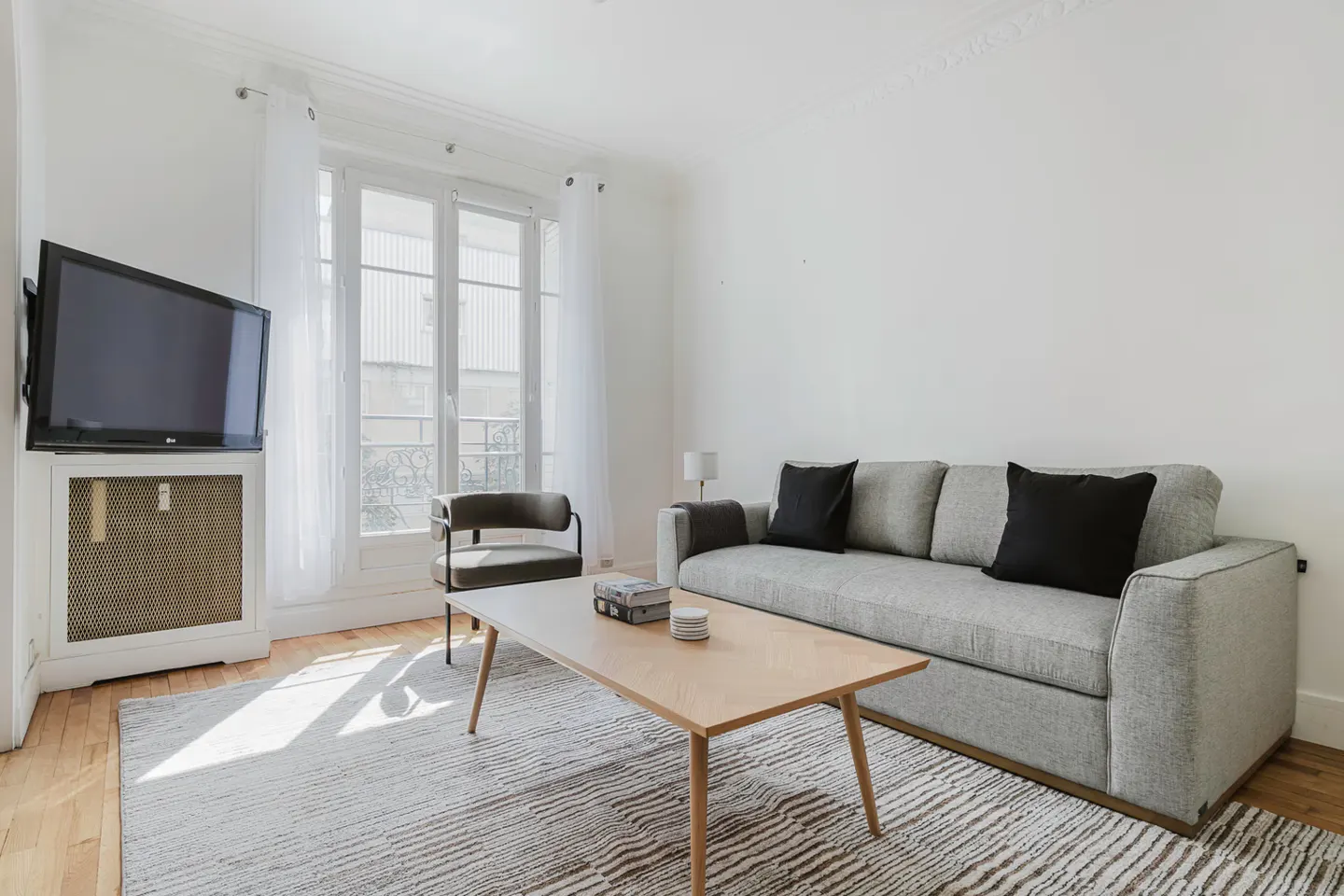 Bright living room with a gray sofa, black pillows, wood coffee table, and a TV mounted above a radiator cover. Sunlight streams through a window.