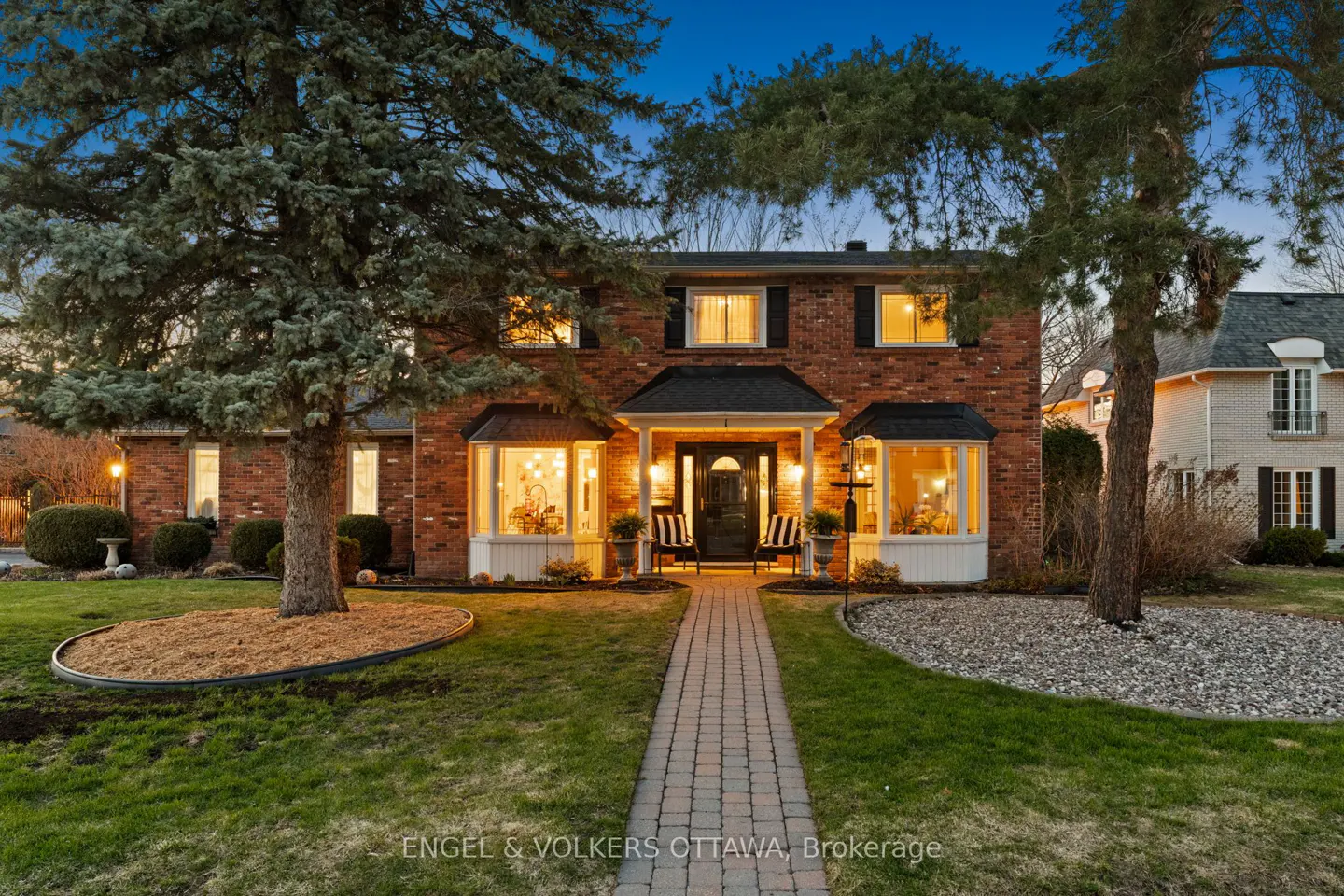 Two-story brick house with a brick walkway, lawn, and trees at dusk. The house has a black door and black shutters. Interior lights are on.