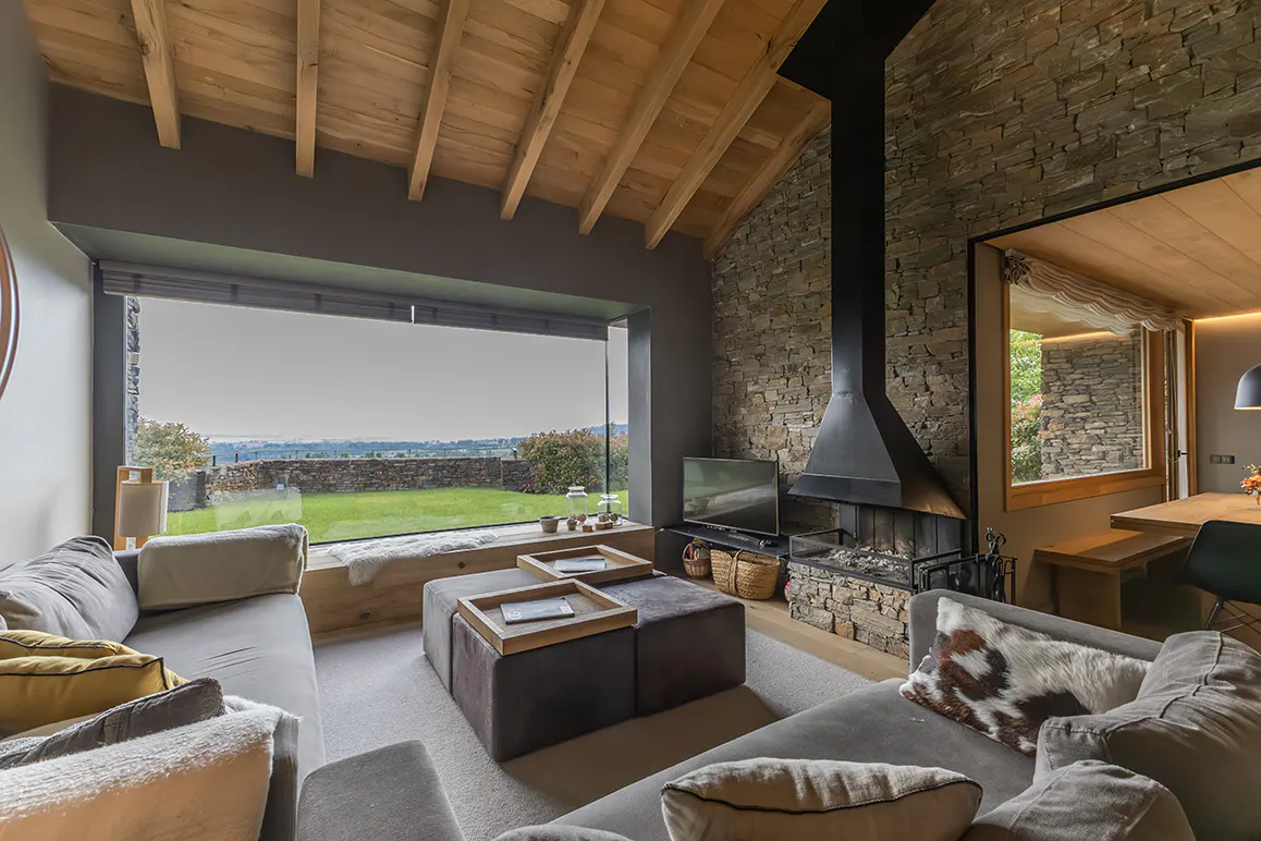 Modern living room with a stone fireplace, vaulted wood ceiling, and a large window overlooking a green lawn.