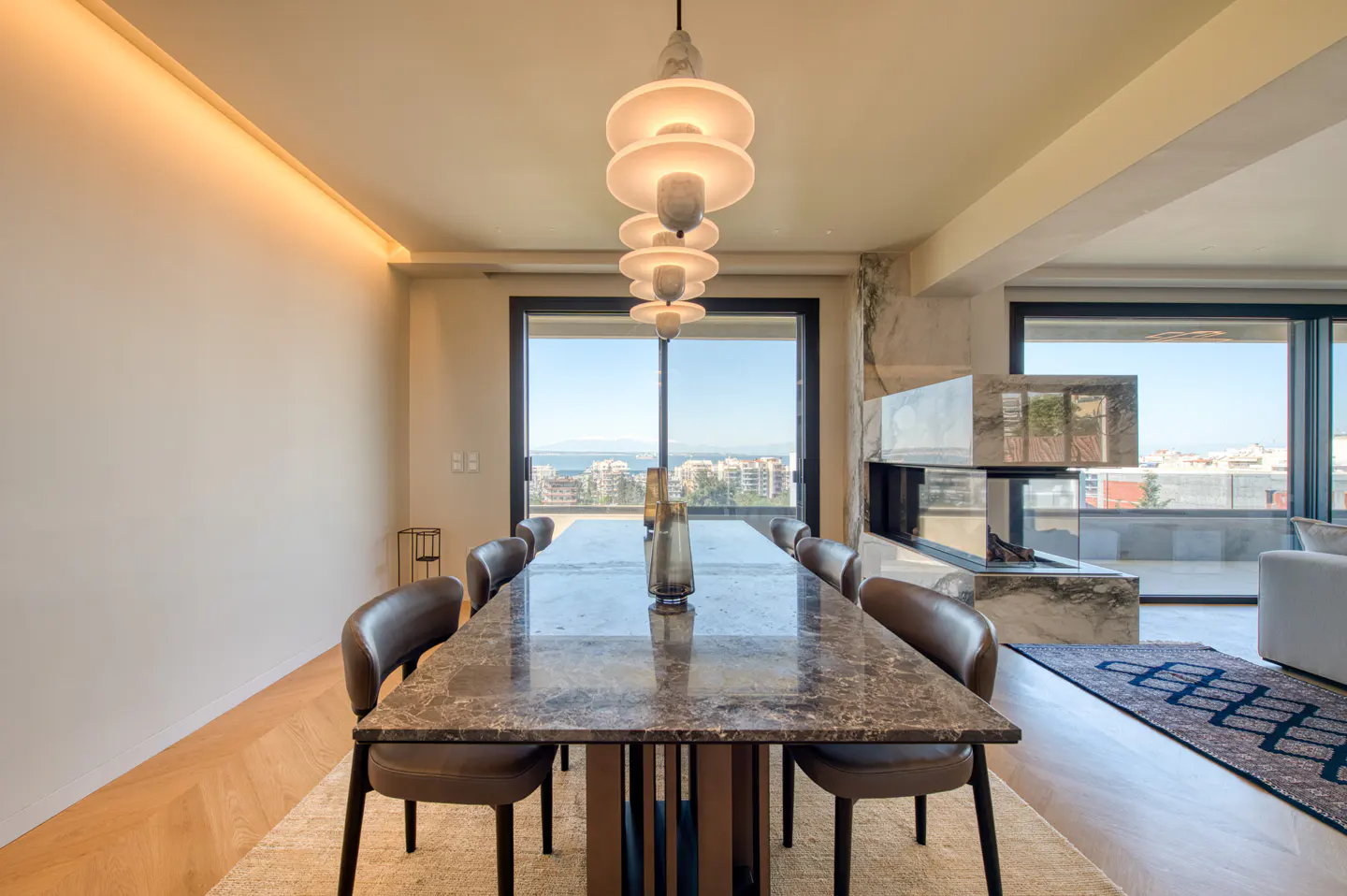 A modern dining room with a marble table, brown leather chairs, and a tiered light fixture. A fireplace and city view are in the background.