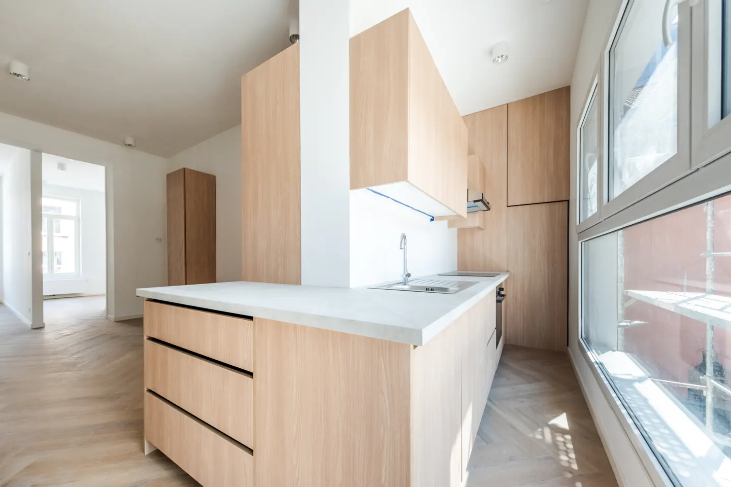 Bright kitchen with light wood cabinets, white countertops, and large windows. Herringbone wood floors and an open doorway to another room.