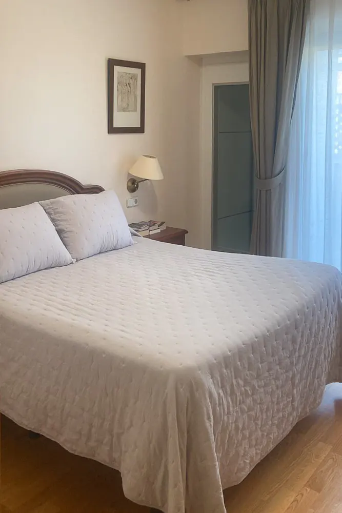 A bedroom with a large bed featuring a white quilted cover, a wall-mounted lamp, framed art, and curtains over a window.
