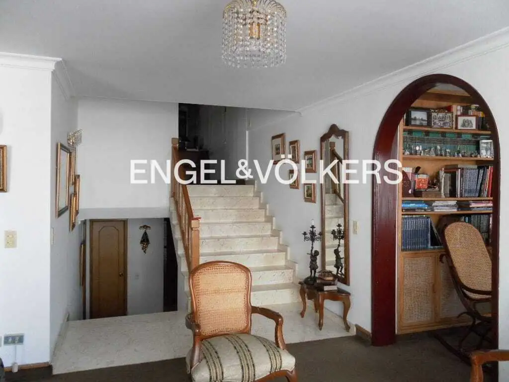 A bright foyer with white walls, marble stairs, and a crystal chandelier. A dark wood archway leads to a bookcase. A chair sits in the foreground.