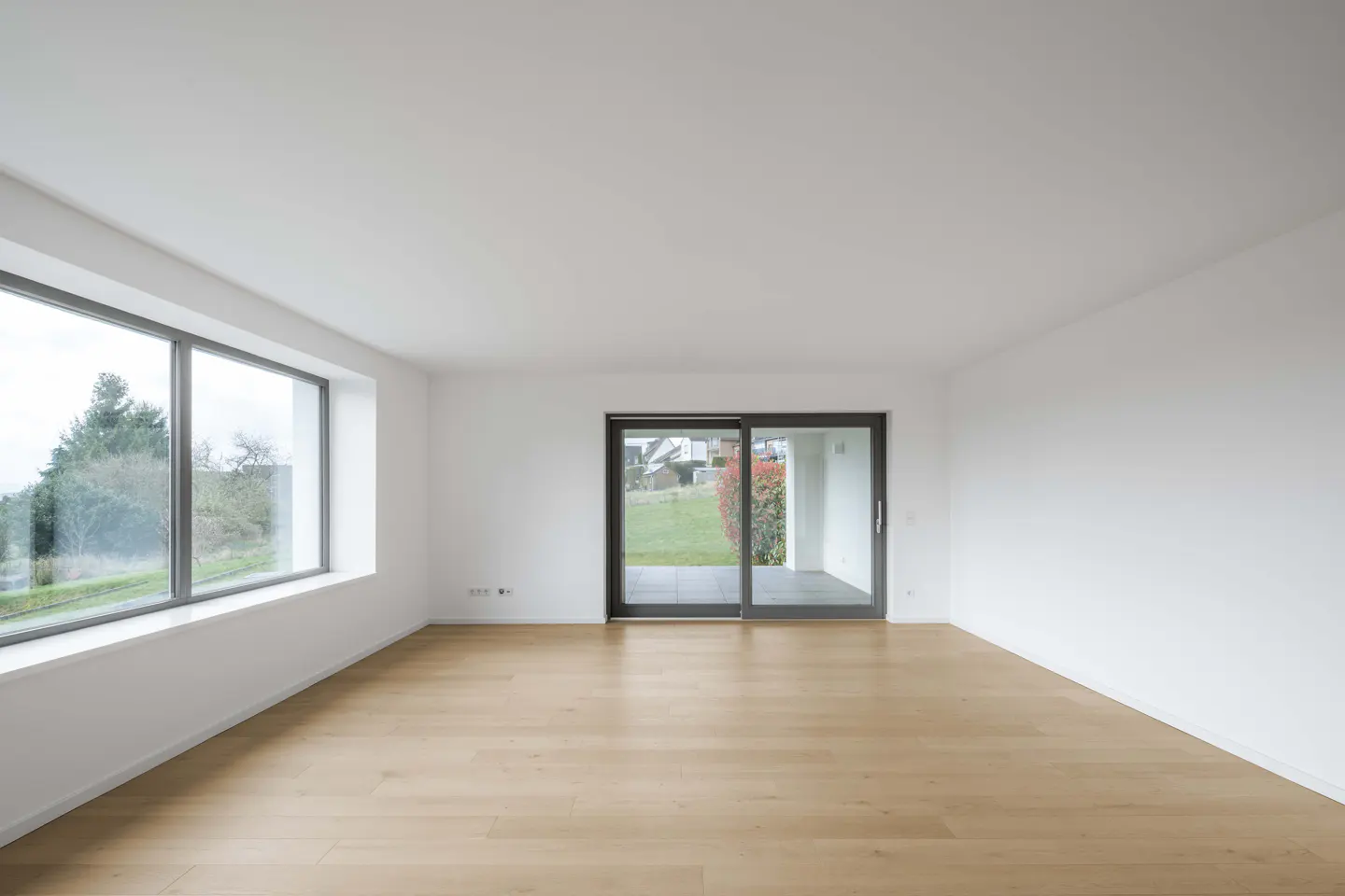 Bright, empty room with light wood floors, white walls, and a large window overlooking trees and a sliding glass door to a patio.