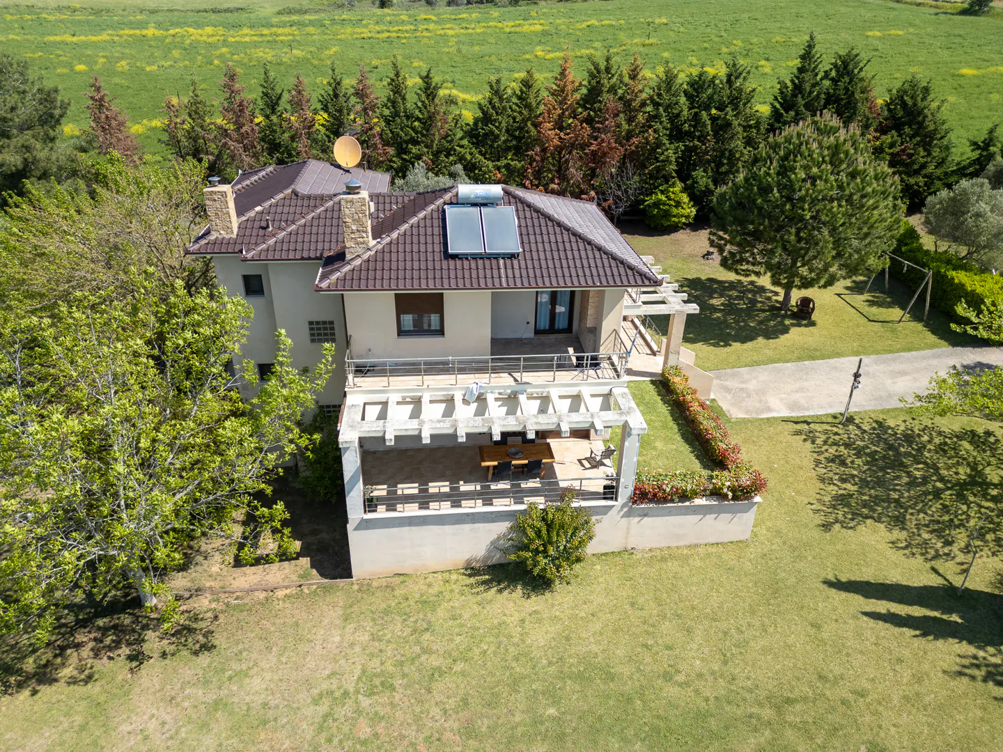 Aerial view of a two-story house with a brown tile roof, solar panels, and a stone chimney, surrounded by green grass and trees.