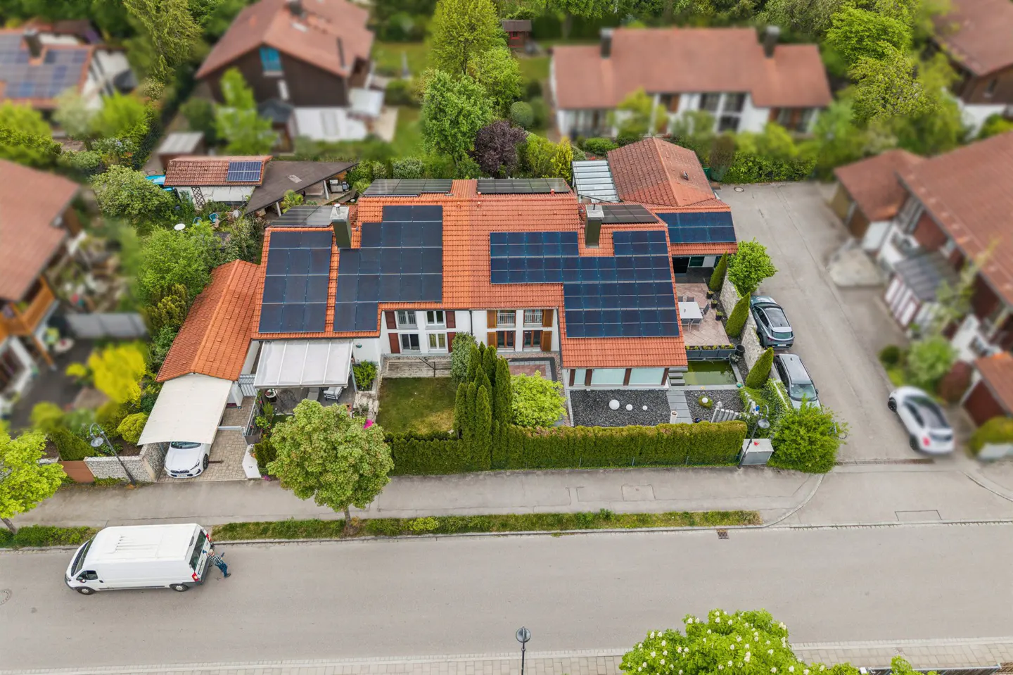Aerial view of a house with solar panels on a red tile roof, surrounded by green trees and other houses in a suburban neighborhood. A white van is parked on the street.