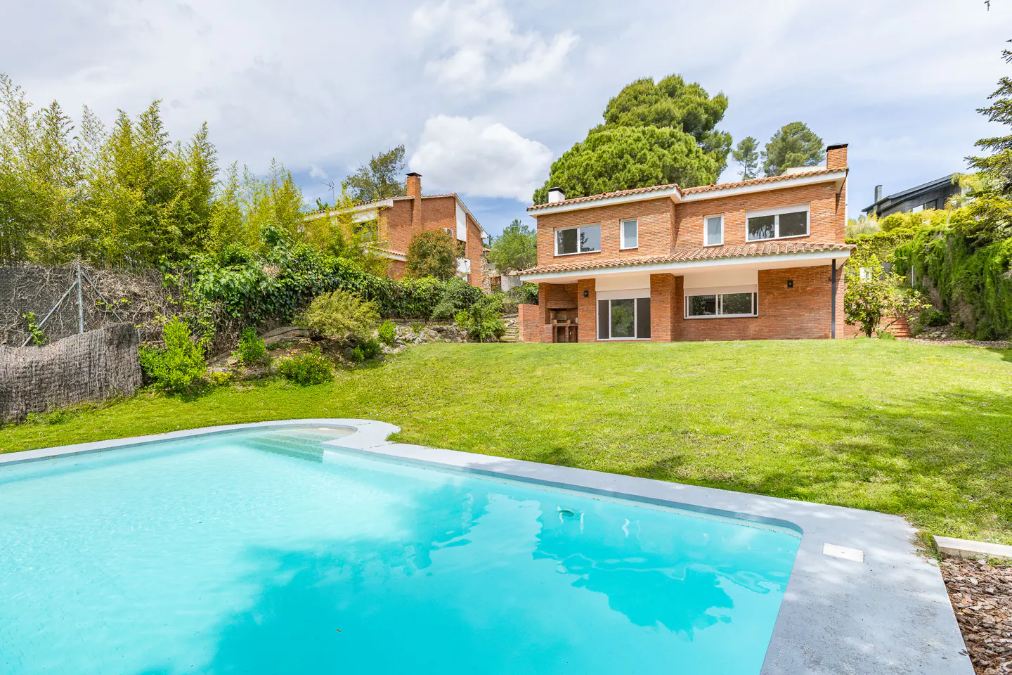 A backyard view of a red brick house with a turquoise pool, green lawn, and trees under a cloudy sky.