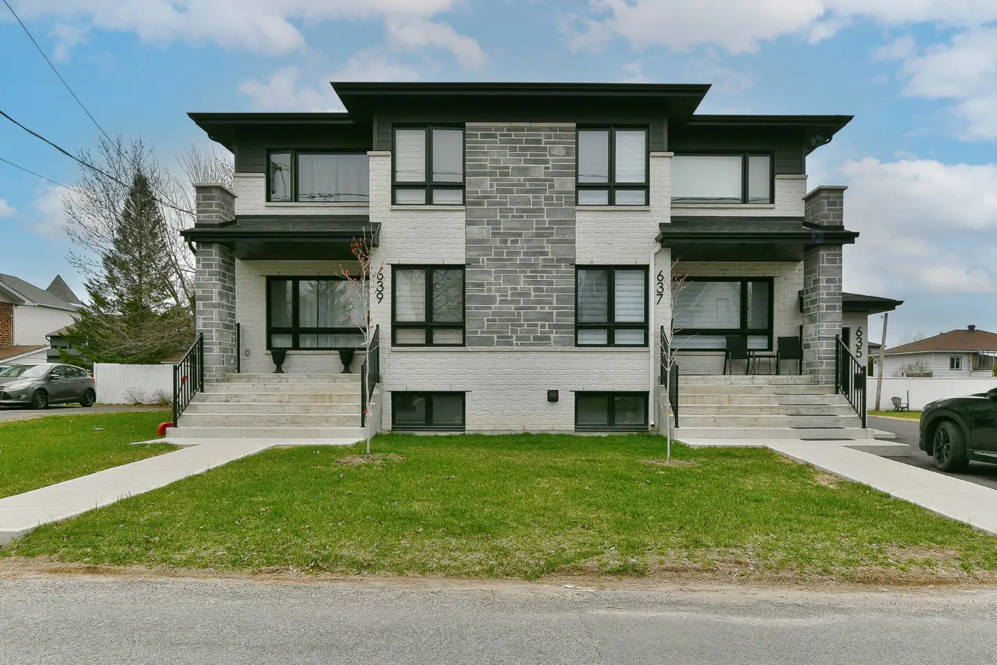 Two-story duplex with white brick and gray stone facade, black trim, and concrete steps leading to each entrance. Green lawn in front.