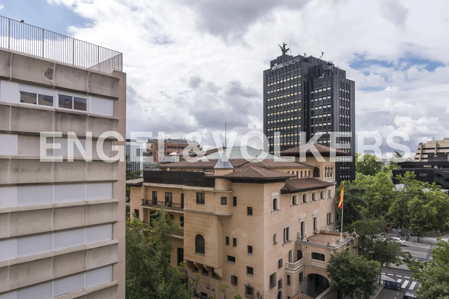 View of a tan building with a red tile roof, a black skyscraper, and green trees under a cloudy sky.
