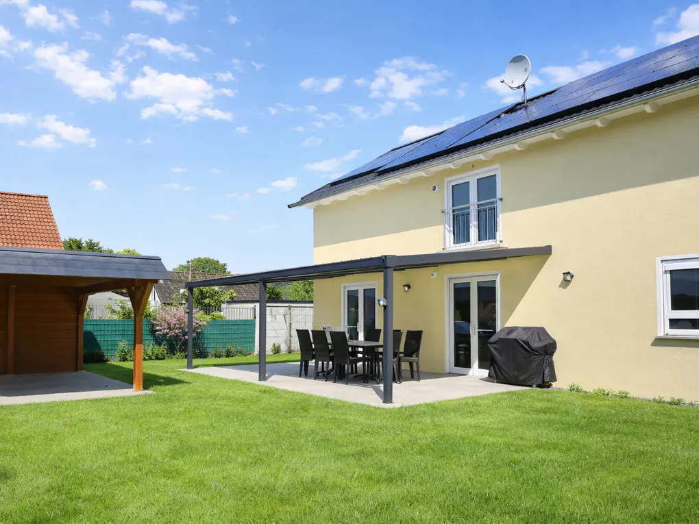 Backyard of a yellow two-story house with solar panels, a patio with a table and chairs, and a wooden carport.