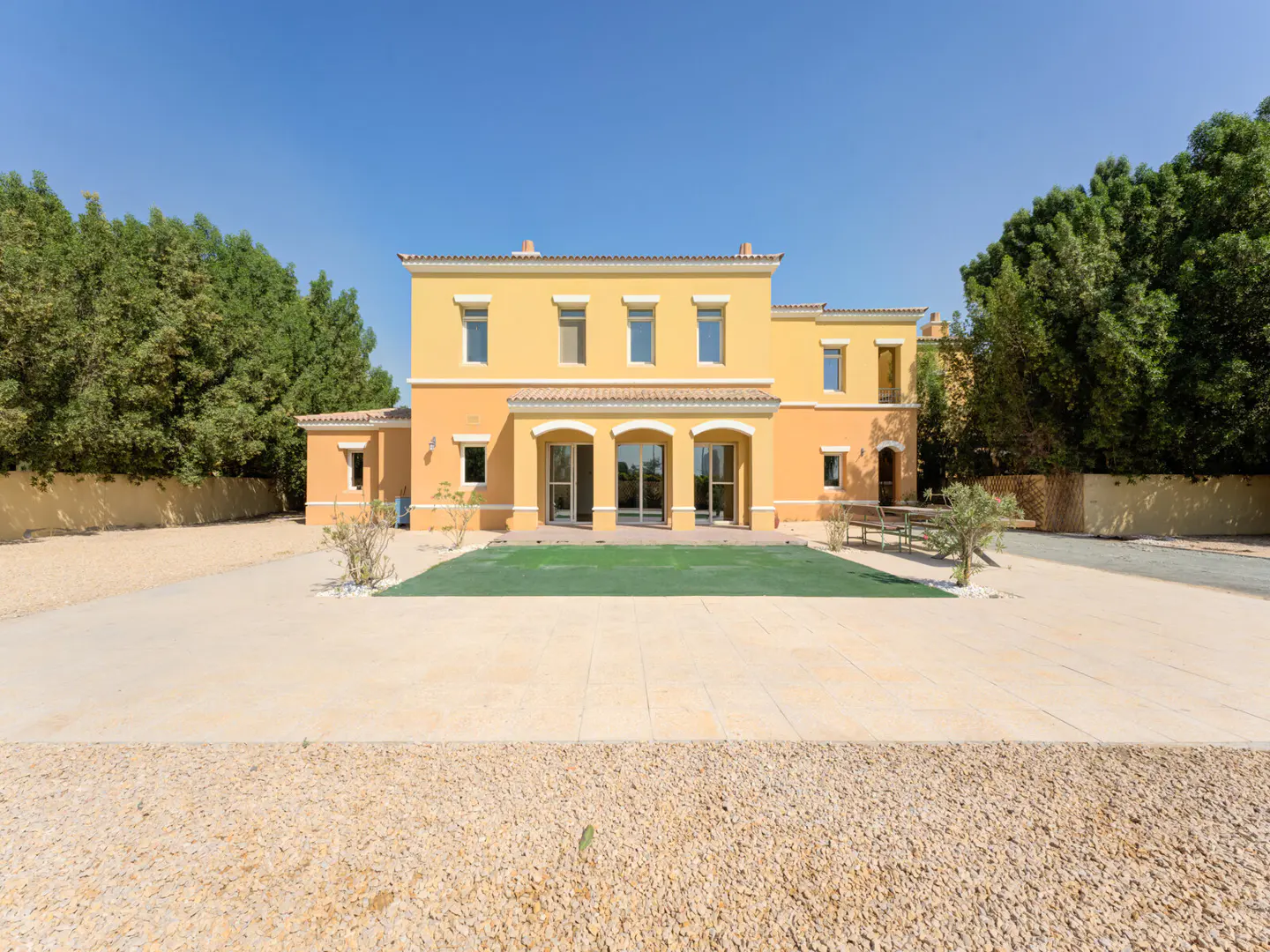 Two-story yellow house with arched patio doors, surrounded by green trees and a gravel driveway under a clear blue sky.