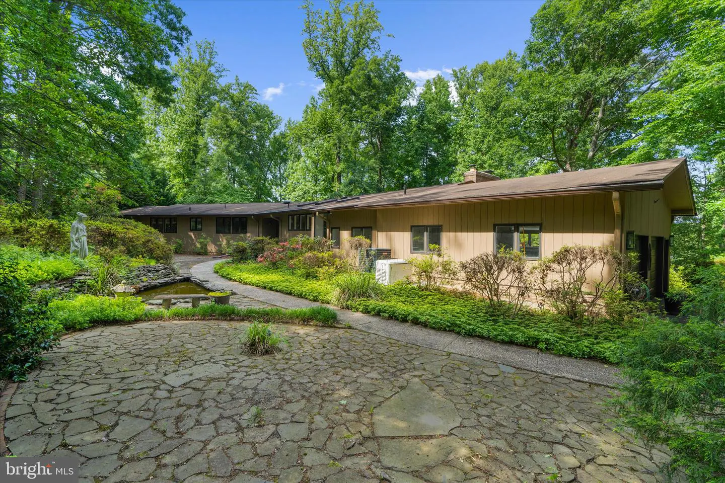Tan ranch-style house with a stone patio, garden pond, and statue, surrounded by lush green trees under a blue sky.