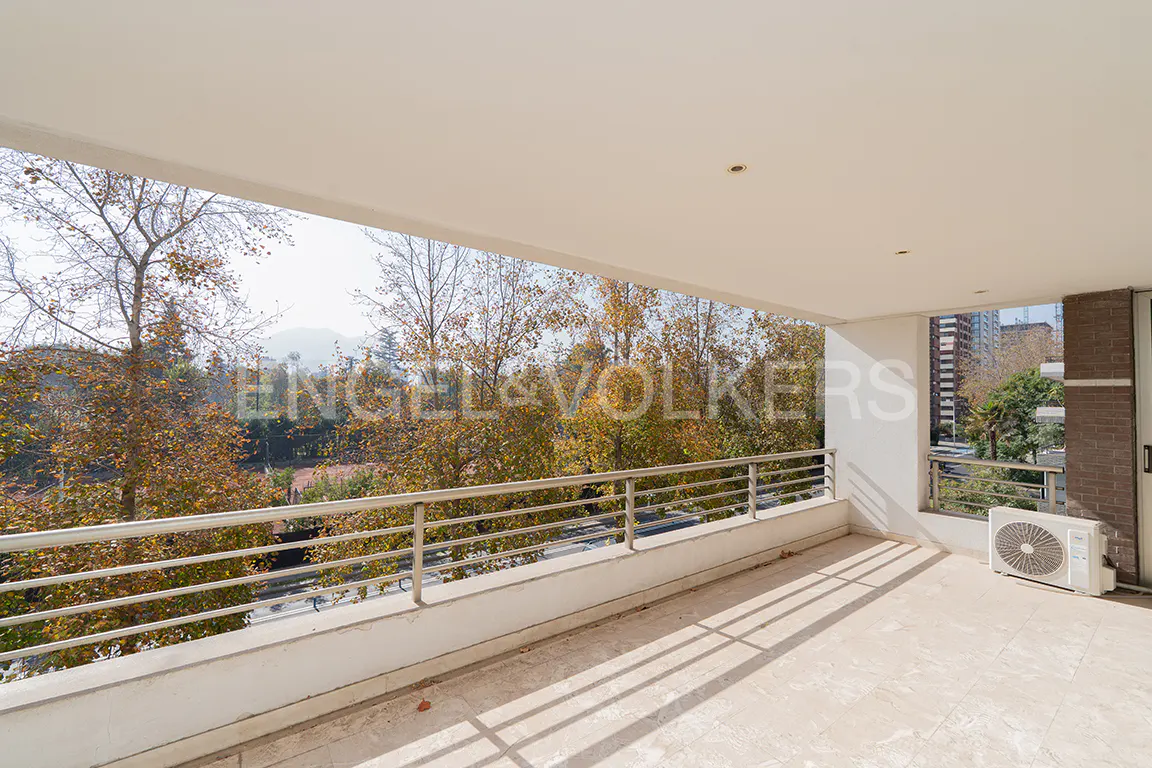 A bright, covered balcony with a beige tile floor and a metal railing overlooking trees and a distant city. An AC unit sits in the corner.