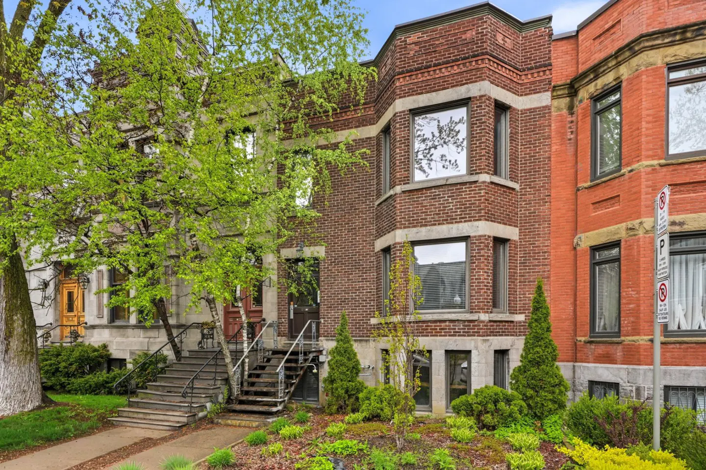 Exterior view of three brick townhouses with stairs leading to front doors and green landscaping.