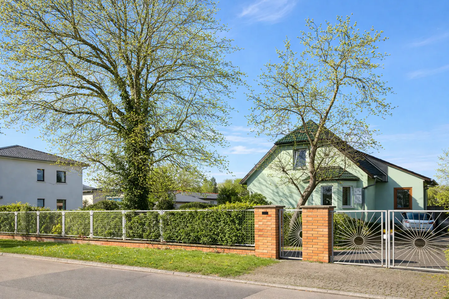 Exterior view of a light green house with a dark roof, a metal gate, and a large tree in the yard.