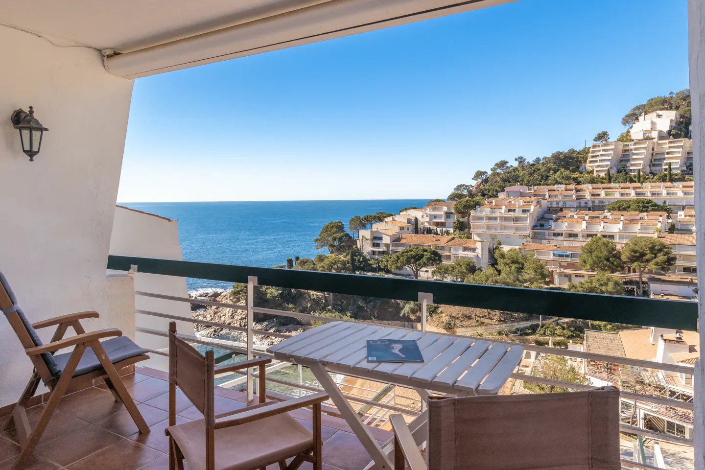 Balcony view with chairs, table, and ocean. Buildings on a hill in the background. Blue sky.