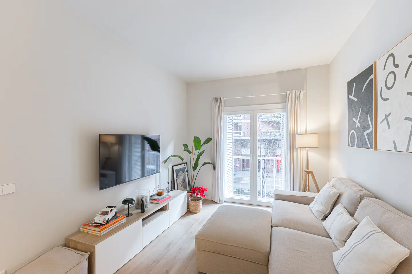 Bright living room with a beige sectional sofa, TV, and white media console. Artwork hangs on the wall, and natural light streams through the balcony doors.