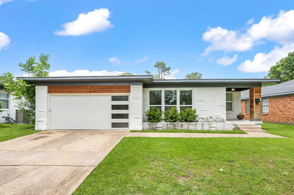 A white brick house with a flat roof, a white garage door with windows, and green bushes in front.