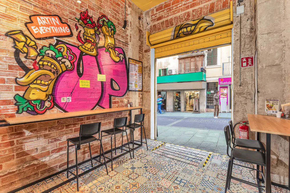 Restaurant interior with brick walls, colorful graffiti, and black stools. An open yellow door leads to a street scene.