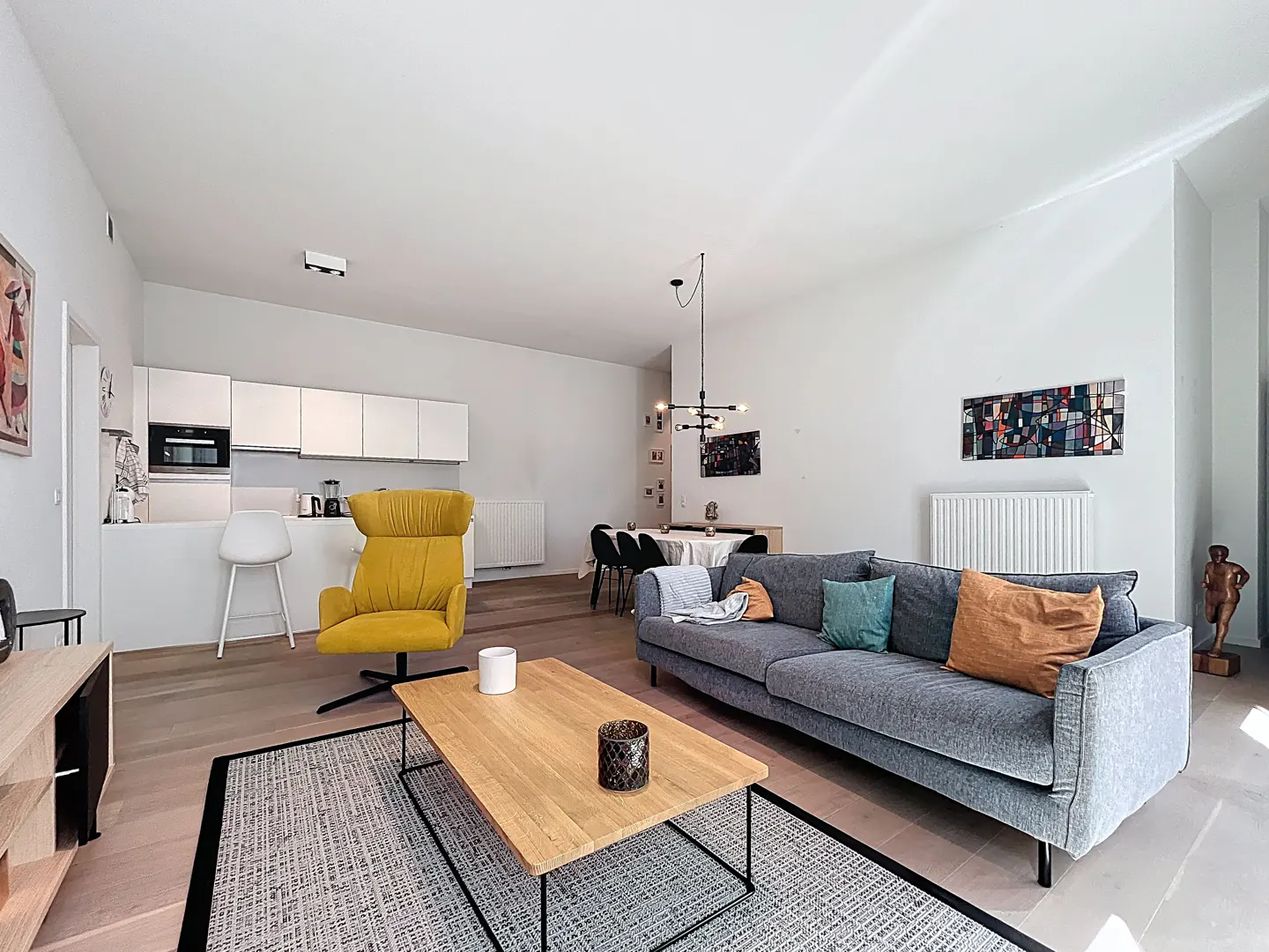 Bright, open-plan living space with a gray sofa, yellow chair, and wooden coffee table on a patterned rug. White kitchen in the background.