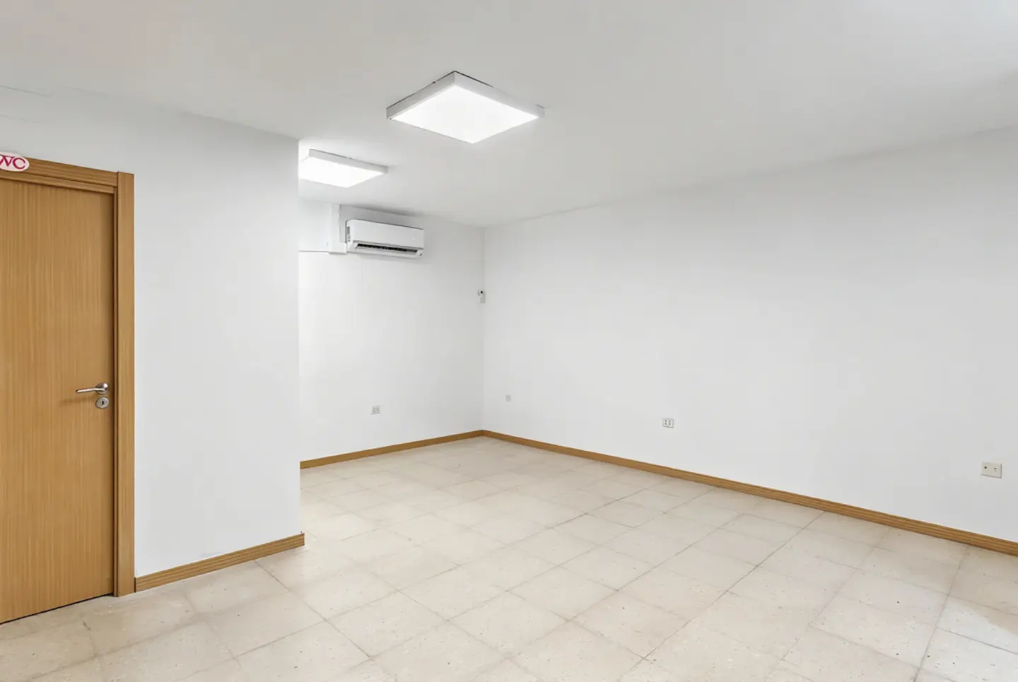 Bright, empty room with white walls, tiled floor, and wood trim. A wood door with a WC sign is on the left. An air conditioner is mounted high on the wall.
