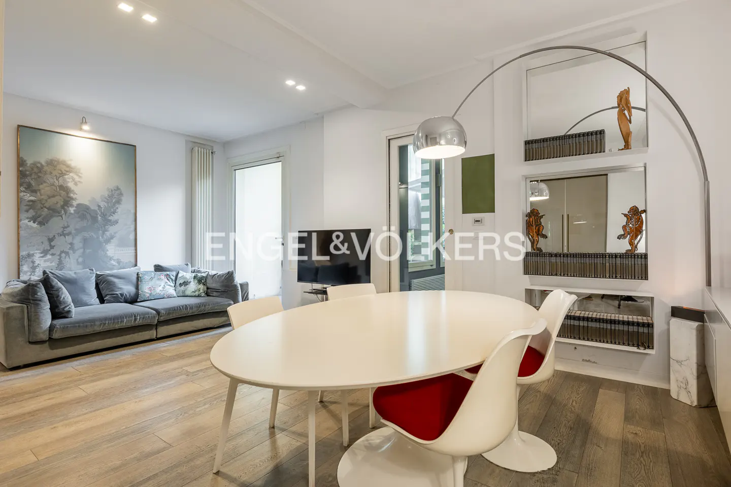 Bright living room with a gray sofa, a large painting, and a white oval table with red and white chairs. A curved floor lamp stands nearby.
