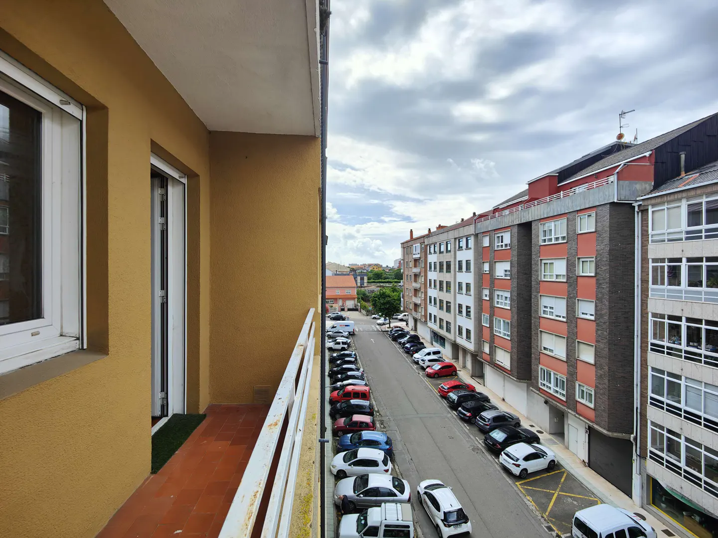View from a balcony with white railings, looking out onto a street lined with parked cars and apartment buildings under a cloudy sky.