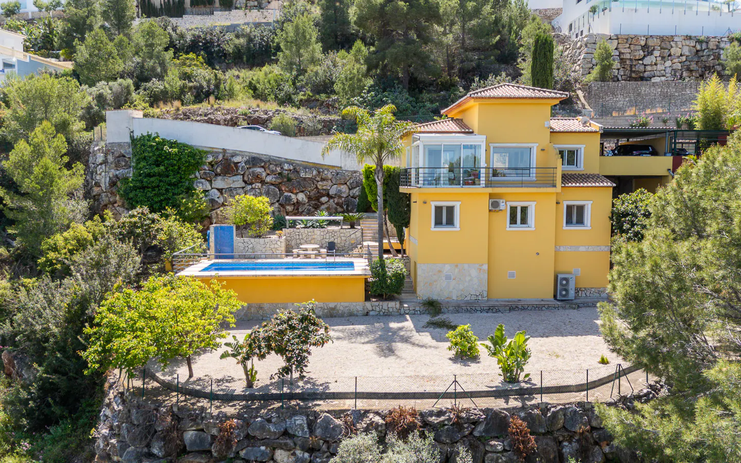 A yellow two-story house with a pool sits on a hillside with trees and stone walls.
