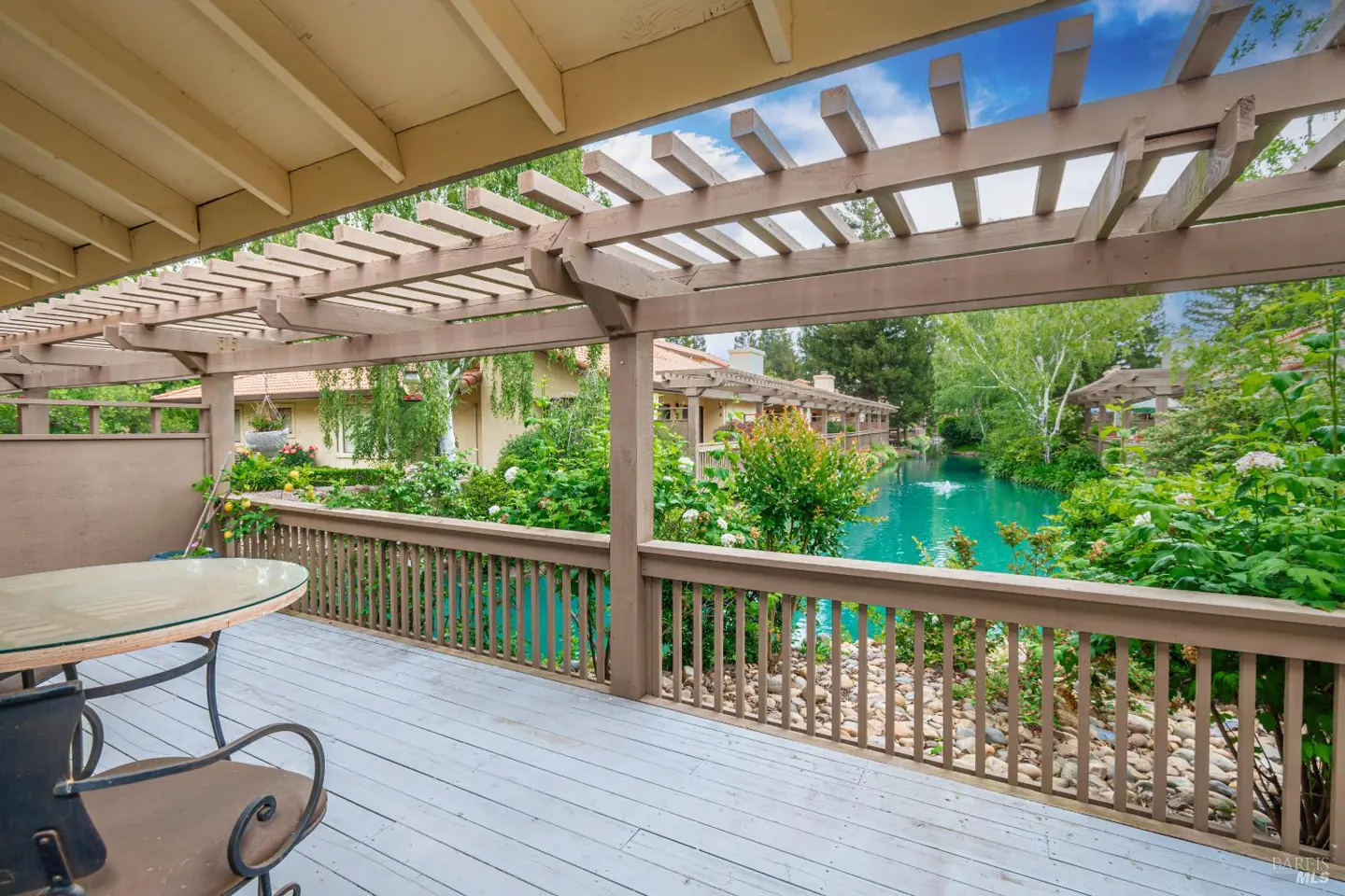 Outdoor patio with a table and chairs overlooking a turquoise pond with lush greenery and a pergola overhead.