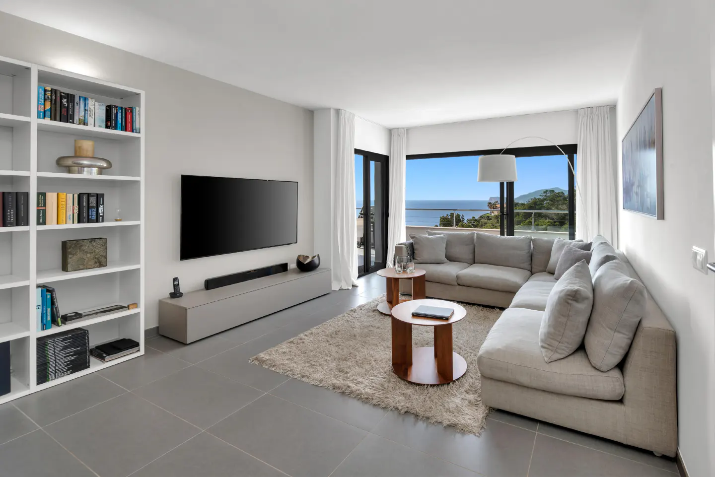 Bright living room with gray tile floor, white walls, and a large gray sectional sofa facing a TV. Ocean view through sliding glass doors.