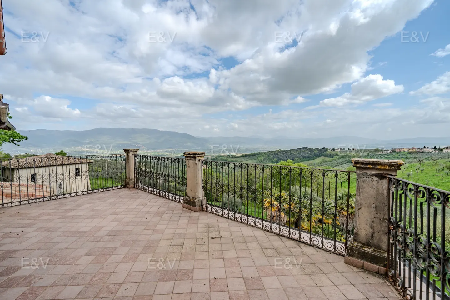 Terracotta-tiled terrace with wrought iron railing overlooking a green valley and distant mountains under a cloudy sky.