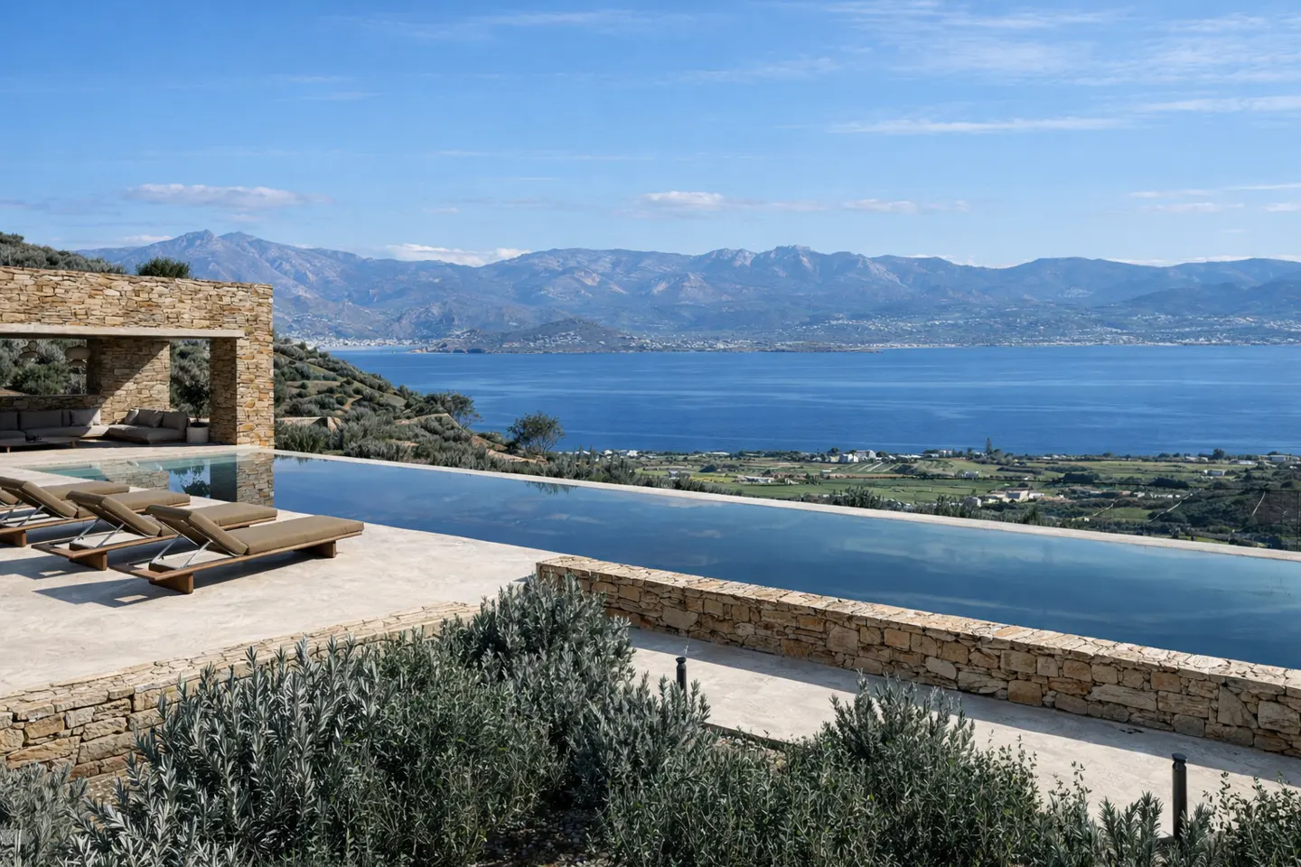 Infinity pool overlooking the ocean. Stone patio with lounge chairs. Mountains and blue sky in the background.