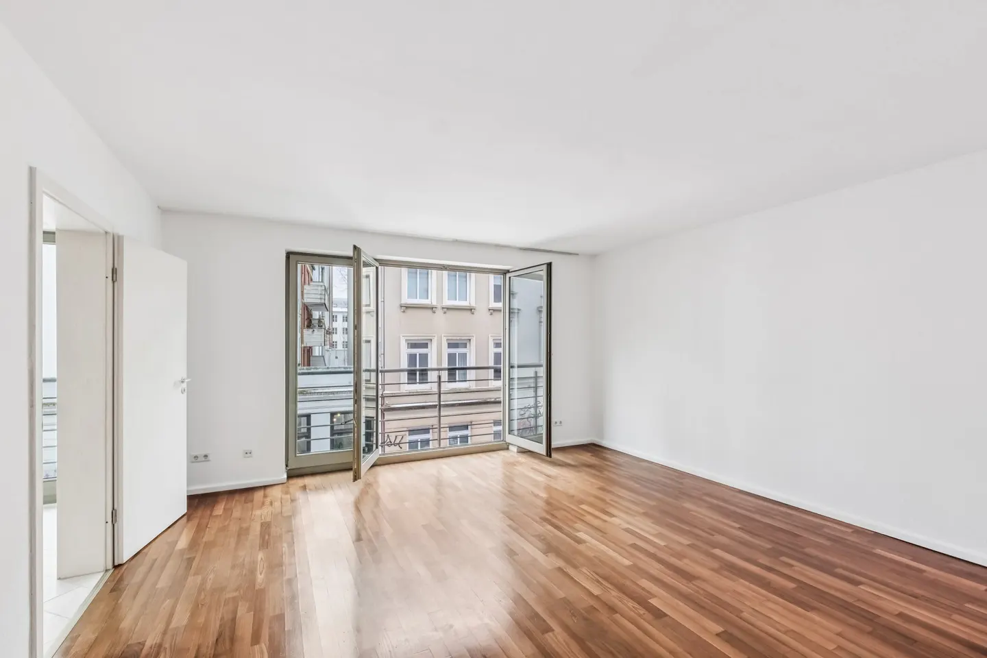 Bright, empty room with hardwood floors, white walls, and open balcony doors showing a city view.