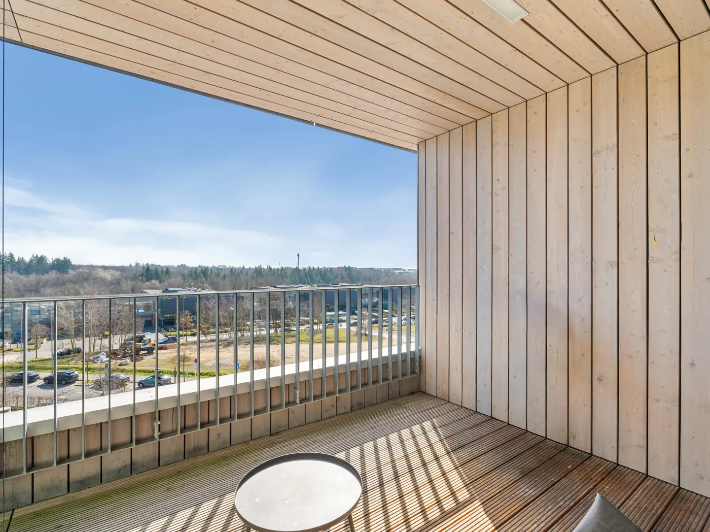 Balcony with wood floor, metal railing, and wood walls. A small table sits on the deck, with a view of trees and a blue sky.