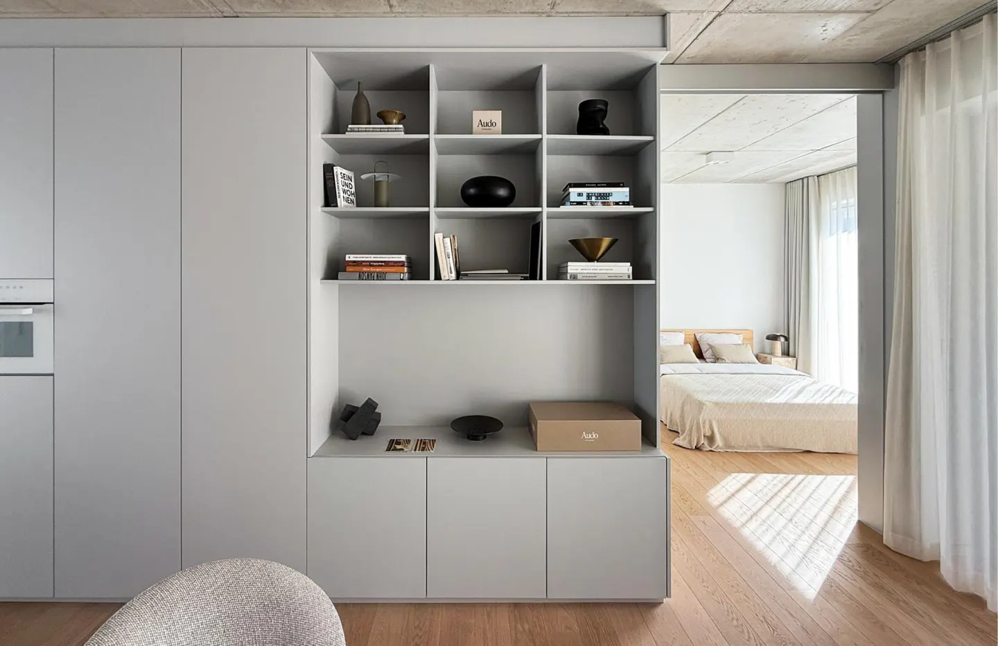 Modern apartment interior with gray built-in shelving unit filled with books and decorative objects, and a view into a bedroom.