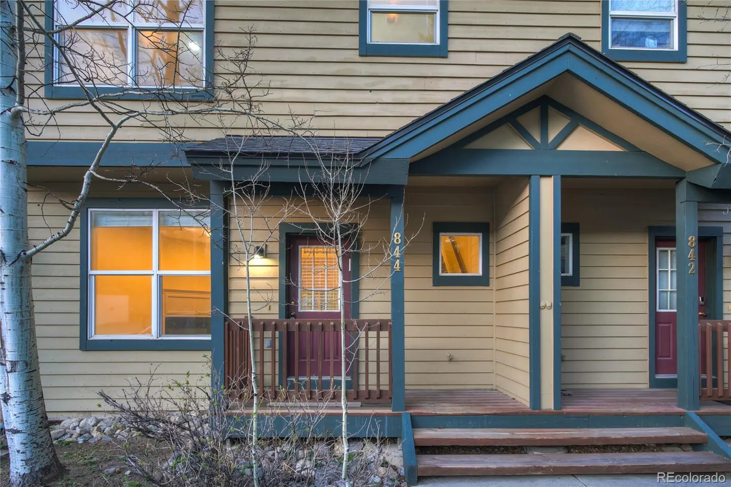 Tan townhouse exterior with blue trim, a small porch, and a red door. Windows glow with warm light.