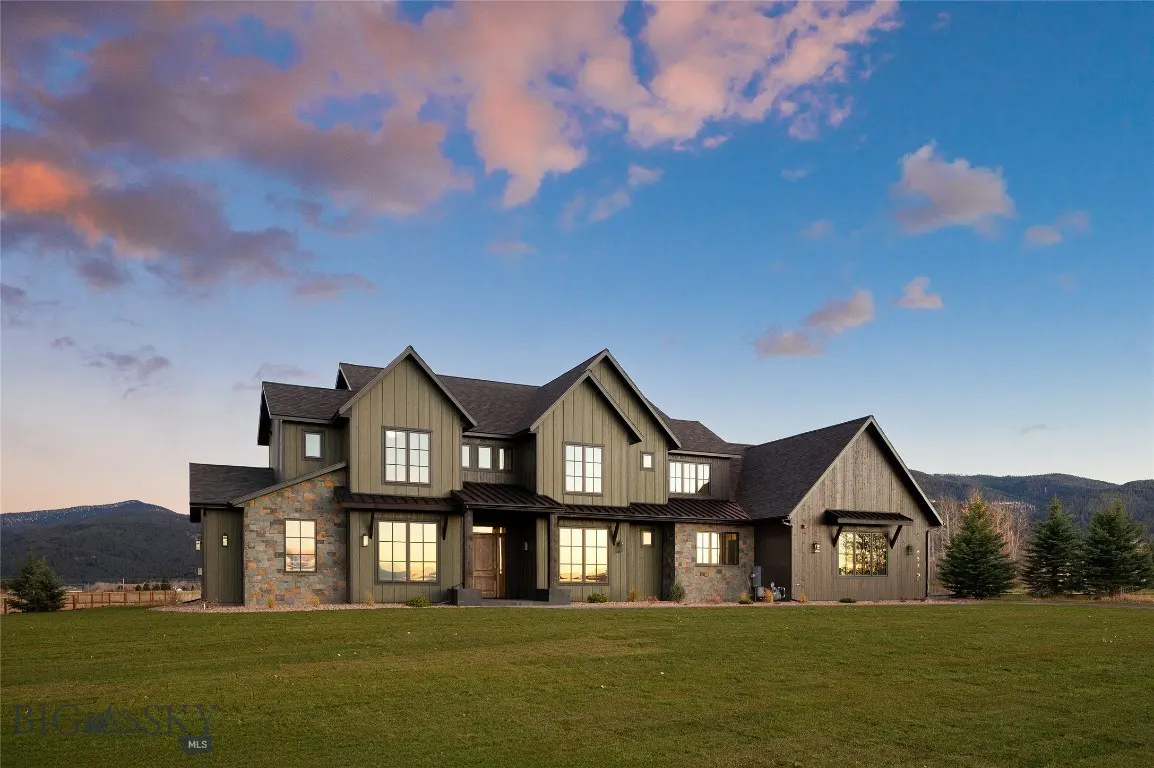 A large, two-story house with green siding and a stone facade sits on a green lawn under a blue and pink sky.